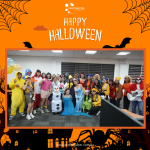 A group of people in Halloween costumes poses indoors with a "Happy Halloween" sign above them. The setting features thematic decorations.