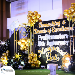 Decorated stage with gold and black balloons and a sign reading "Unmasking a Decade of Excellence: Profitmaster's 10th Anniversary Masquerade Ball.