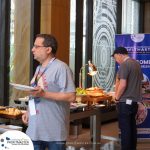 Two men are standing near a buffet table at a conference; one is holding a plate of food, and a banner with the text "Profit Master" is partially visible in the background.