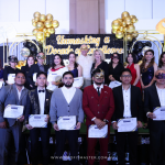 A group of people in formal attire pose for a photo on stage holding certificates. The backdrop reads "Unmasking a Decade of Excellence" and is decorated with balloons and masks.