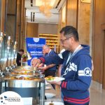 A man serves himself food from a buffet at a conference event. Banners in the background display "Profitmaster Global Outsourcing." Other attendees are visible in the background.