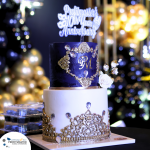 A two-tiered cake with blue and white layers, ornate gold decorations, and a "Profitmasters 10th Year Anniversary" topper. Golden balloons and lights are blurred in the background.