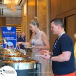 Two people serve themselves from a buffet at a conference event. A banner in the background displays "Profitmaster." The setting appears to be a hotel or conference center.