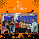 A group of people in costumes hold number cards in an office decorated for Halloween with spider web and bat motifs. Text at the top reads "Happy Halloween.