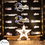 A backdrop featuring multiple company logos, a "Profitmaster Global Outsourcing" anniversary message, and a large illuminated star decoration at the bottom.