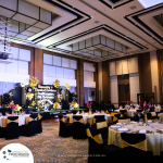 A decorated event hall with round tables set for a formal gathering. There are black and gold balloon arrangements and a stage with a sign that reads, "Congratulations to a happy retirement.