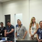 A group of people stands in a conference room, with a man speaking into a microphone. A screen displays "Welcome, Speakers and Delegates" along with conference details in the background.