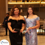 Two women in elegant dresses stand side by side at an indoor event. The woman on the left wears a black dress, and the woman on the right wears a light blue dress.