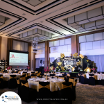 A banquet hall is set up with round tables and chairs for an event. A stage in the background features a decorative backdrop, and projection screens display event details. The room is well-lit and elegant.