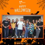 A group of people in costumes and casual wear stand together, smiling, in a Halloween-themed setting with an orange "Happy Halloween" banner above.