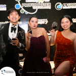 Three people in formal attire stand in front of a branded backdrop, holding wine glasses and smiling at an event.