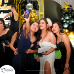 A group of women in formal attire take a selfie at an event. They stand in front of a backdrop decorated with gold and black balloons.