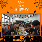 A group of people sit at a long table enjoying a meal, with Halloween decorations surrounding them. A "Happy Halloween" banner is displayed above.