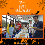 Group of people at a Halloween-themed party, standing in a line, serving themselves food from a table with various dishes. Orange and black decorations are visible in the background.