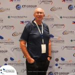 A man in a navy polo shirt and glasses stands in front of a branded backdrop at a ProfitMaster event.