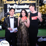 Three people stand posed, smiling and holding envelopes at an event celebrating Profitmaster's 10th Anniversary Masquerade. The backdrop features the event's name with gold and black decorations.