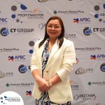 A woman wearing glasses and a white blazer stands in front of a backdrop covered with various company logos, including ProfitMaster, CT Group, Assure Global Plus, APBC, CLARK, and ANZCHAM Philippines.