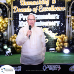 A man in a traditional shirt speaks into a microphone in front of a backdrop with "Unmasking a Decade of Excellence, Profitmaster's 10th Anniversary" text. Balloons and lights decorate the scene.