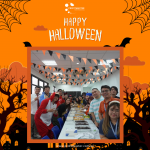 A group of people in Halloween costumes stand around a table with food. The background is decorated with Halloween-themed designs.