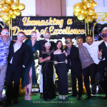 A group of people wearing masks pose in front of a backdrop reading "Unmasking a Decade of Excellence" with decorative balloons and gold stars.