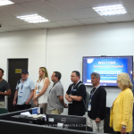 A group of people standing in line in an office setting. A screen on the wall displays the text, "WELCOME, Speakers and Delegates! PROFITMASTER." There is a banner on the left side of the image.