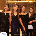 Four women in black evening dresses stand together, posing for a photo at a formal event with a "Profit Master" sign in the background.