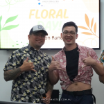 Three men in floral shirts stand smiling in front of a "Floral Day" sign, with one man holding a microphone.