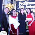 Four people dressed formally pose for a photo at an event. One of the women is holding a certificate or award. Balloons and a banner in the background indicate it is a 10th anniversary celebration.