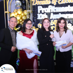 Four people stand together, smiling, at the Profitmaster 10th Anniversary event. A sign behind them reads "Decade of excellence, Profitmaster 10th Anniversary." They are holding white envelopes.