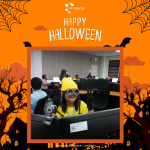 Office with employees in Halloween costumes, one person in the foreground wearing a yellow outfit and goggles, set against a festive orange themed backdrop with "Happy Halloween" text.