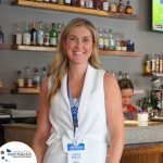 A woman wearing a white outfit and a name badge stands smiling in front of a bar counter with bottles and a coffee machine in the background. Another person is seen working behind the bar.