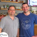 Two men smiling and standing side-by-side in front of a bar with various liquor bottles. One man is wearing a striped shirt and glasses, and the other is wearing a blue t-shirt with a mountain design.