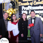 Four people dressed formally stand together at an event celebrating Profitmaster's 10th anniversary. The person on the right is holding a red envelope. Balloons and a banner are in the background.