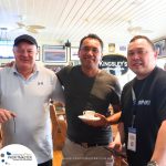 Three men are standing in a casual indoor setting. One is holding a white mug. A PROFITMASTER banner and various framed pictures are visible in the background. All are smiling.