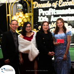 Four people stand together at a formal event in front of a backdrop that reads "Profitmaster 10th Anniversary." All are dressed in formal attire, and one person holds a red envelope.