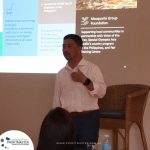 A man stands in front of a projected presentation slide, speaking to an audience. The slide displays information about the Macquarie Group Foundation and community programs in the Philippines.