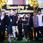 A group of people in formal attire pose for a photo in front of a decorated backdrop with the text "Unmasking a Decade of Excellence," while wearing masquerade masks.