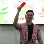 A person in a floral shirt holds up a red card while standing in front of a screen displaying "Floral Day Fashion Friday.