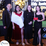 Four people are dressed formally, standing in front of a gold and black decorated backdrop at an event. The women are wearing dresses, and one is holding a red envelope. Logo at the bottom left.