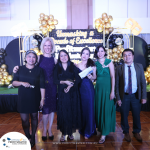 A group of people dressed formally, standing on a wooden floor in front of a backdrop with balloons and the text "Unmasking a Decade of Excellence.