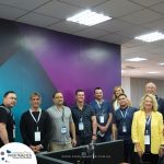 A group of nine people wearing ID badges stands together in an office space with the "Profitmaster Global Outsourcing" logo visible in the foreground.