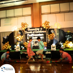 Children in traditional attire perform a Filipino cultural bamboo dance on stage, decorated with a colorful backdrop and balloons, at an event with the theme "Unmasking a Decade of Excellence.