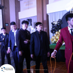 A group of men dressed in formal wear stand in a line at an indoor event. One man in a red suit holds a rope, and balloons are displayed in the background. A banner reads "PROFITMASTER.