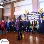 People in formal wear stand on stage at a Profitmaster event celebrating their 10th anniversary, with a backdrop decorated with gold balloons and the event's logo.