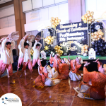 A group of performers in traditional attire dances on a stage with baskets. Behind them, a backdrop reads "Profitmaster's Masquerade Ball." Balloons and decorations adorn the background.