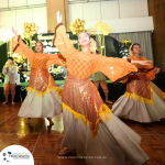 A group of people in traditional costumes perform a dance on a wooden floor with a decorated backdrop and balloons behind them.