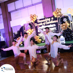 A group of five people performing a dance routine on a wooden floor in front of a stage with decorative balloons and a backdrop that reads "Unmasking a Decade of Excellence.