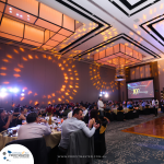 A large group of people sit at decorated tables in a ballroom, attending an event with a presentation on a screen displaying "10th Anniversary." The room is illuminated with warm, decorative lighting.