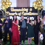 A group of people in formal attire stands in front of a backdrop with the text "Unmasking a Decade of Excellence," surrounded by gold and black balloons and masquerade masks.