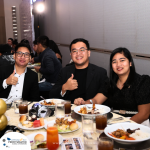 Three people are seated at a dining table with food and beverages, smiling and posing for the camera, with one person giving a thumbs-up.